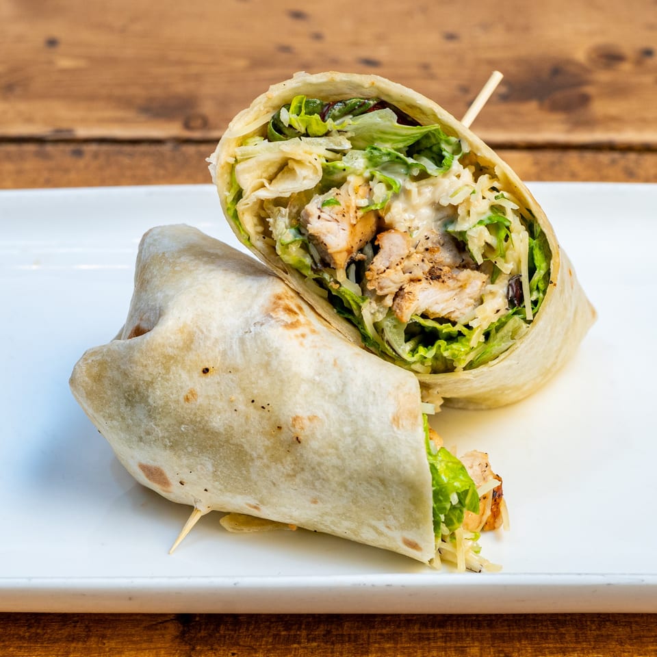 Two halves of a chicken Caesar wrap, filled with grilled chicken, lettuce, and dressing, on a white plate.